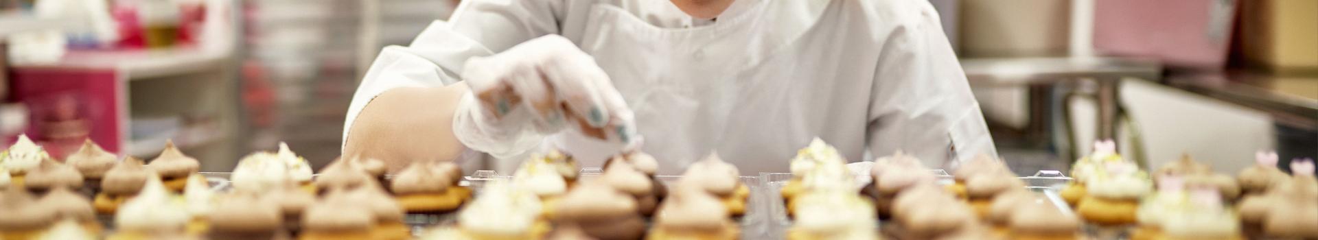 A pastry chef stands behind a table of freshly baked tarts. She leans in close, and, with a gloved hand, put the finishing touches on a dessert. 